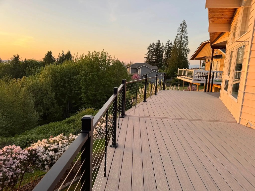 Gray composite deck with black aluminum stainless steel cable railing that overlooks trees and flowers at sunset.